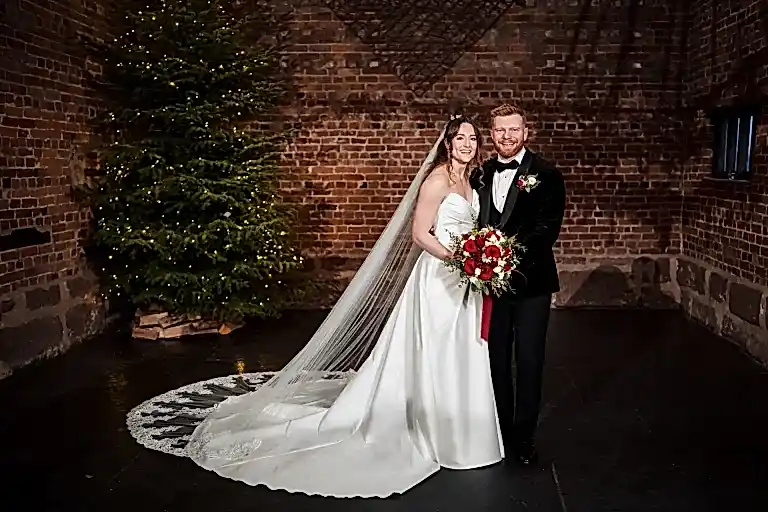 bride and groom pose for their wedding photographer inside Curradine Barns in front of a tall christmas tree