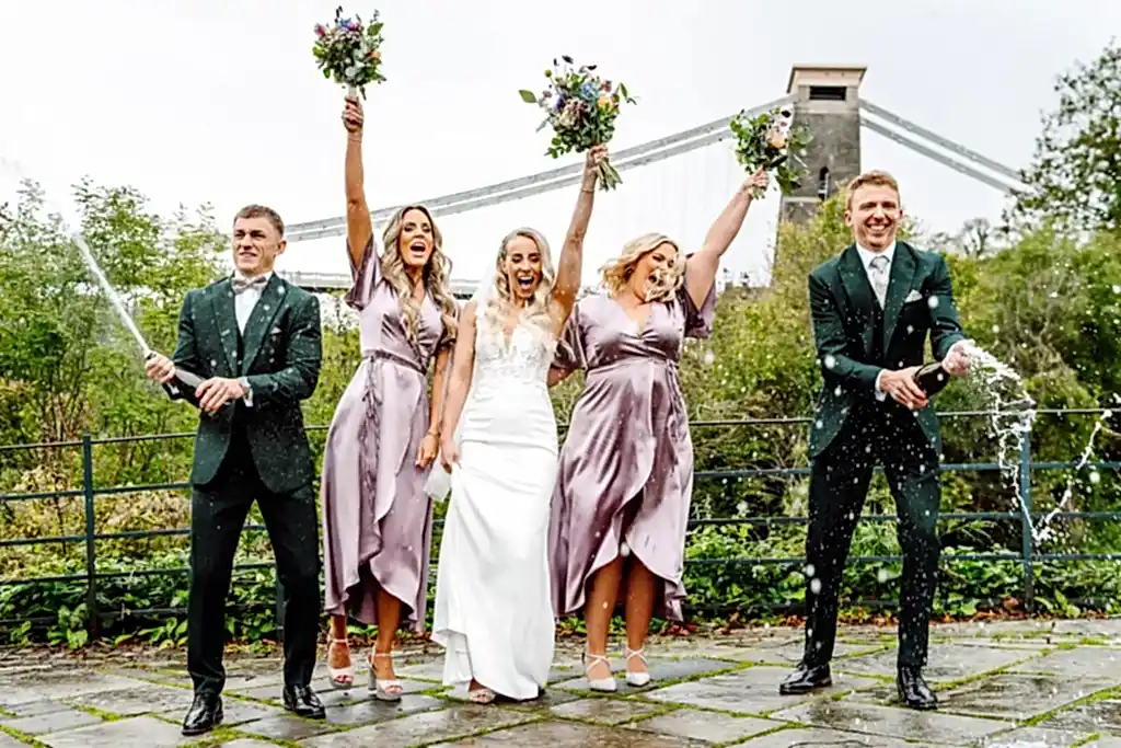 bridal-party-celebrate-champagne-bristol-suspension-bridge a group of people posing for a picture while spraying champagne in front of the Bristol suspension bridge in Clifton