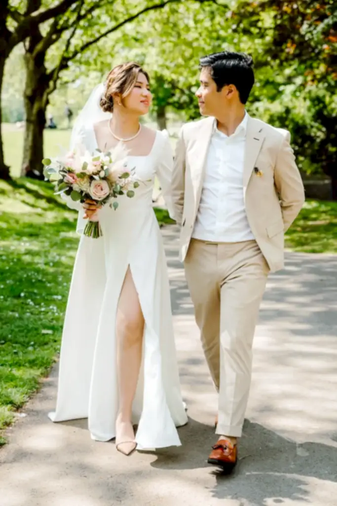 a bride holding flowers and groom in a light cream suit walk through castle park in bristol on a sunny wedding day