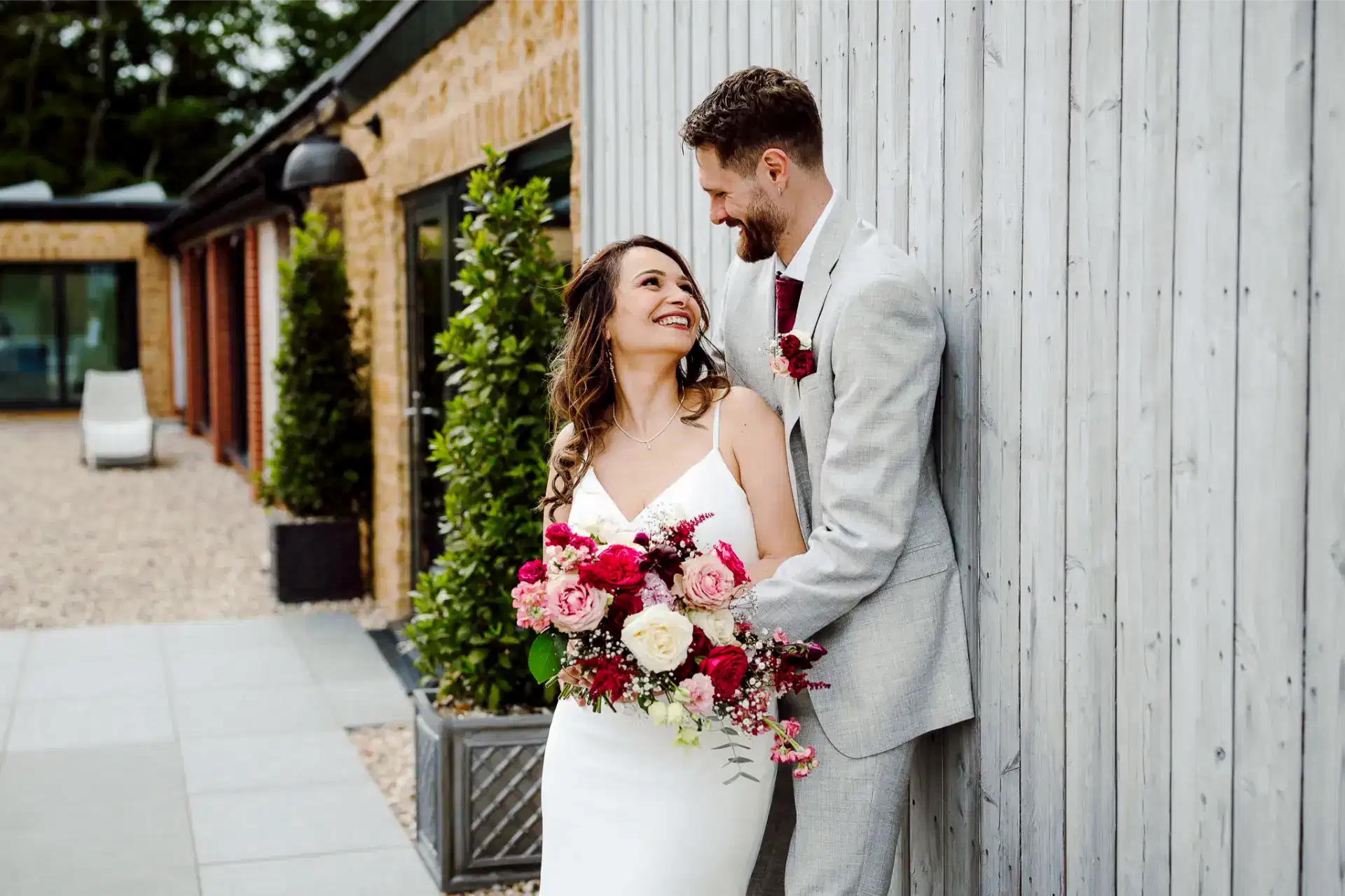 groom leans against wooden wall of a building while his bride leans into him and looks lovingly into his eyes as they pose for their wedding videography