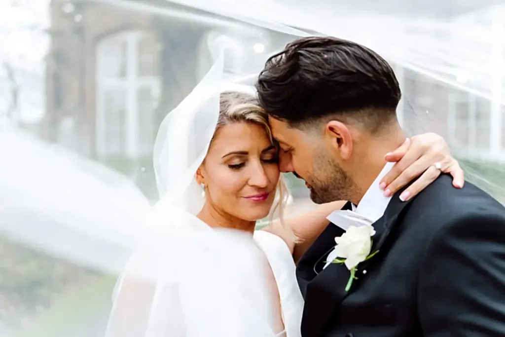 bride-groom-under-veil-st-audries-park bride and groom with their eyes closed embrace under a white veil as it blows in the wind at st audries park wedding venue