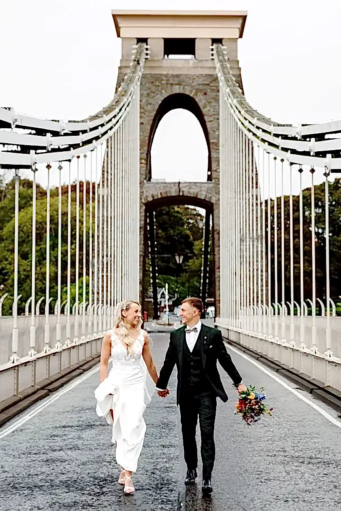 bride-groom-walk-across-clifton-suspension-bridge bride and groom walk down the middle of the clifton suspension bridge looking at one another in this example of bristol wedding videography