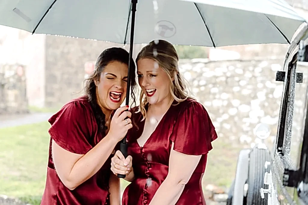 bridesmaids-under-umbrella-raining-clevedon two bridesmaids in red dresses take cover under an umbrella on a rainy wedding day and laugh at the situation