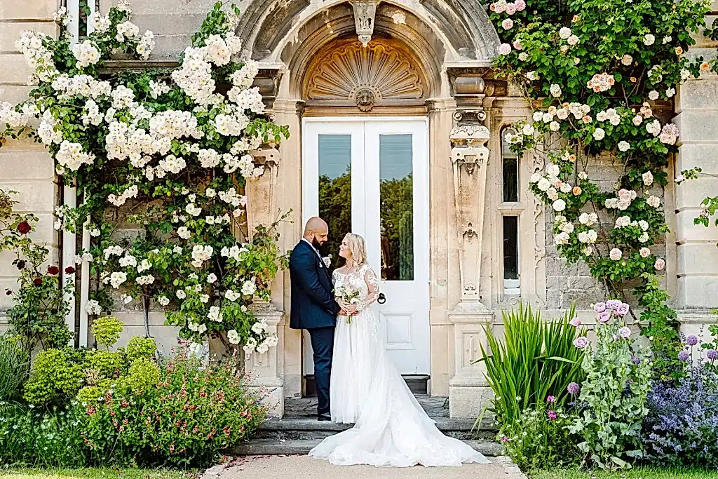 BUT0623-LOWRES-0529 bride and groom stand looking at one another underneath a wall of blooming roses at clevedon hall in somerset