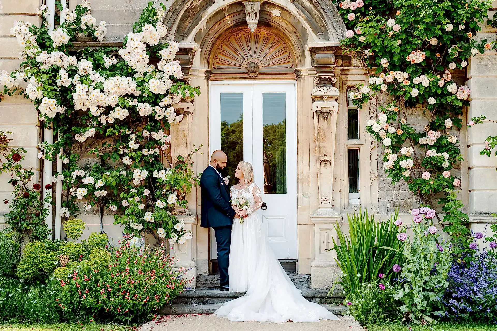 bride and groom stand looking at one another underneath a wall of blooming roses at clevedon hall in somerset