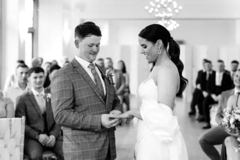 a man in a suit and woman in a white dress exchange wedding rings as their ceremony is reliably live streamed by white villa weddings