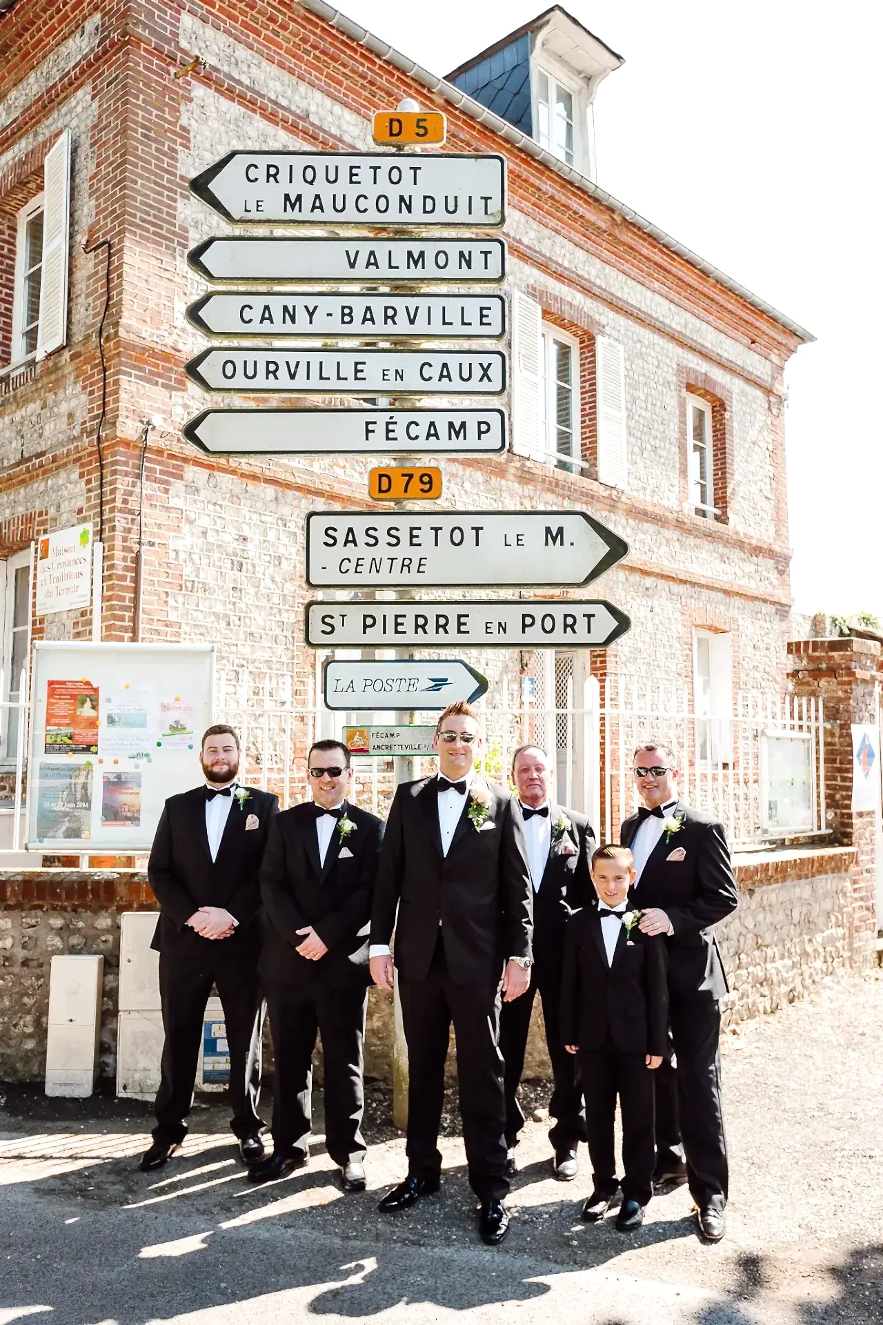 a group of groomsmen in black suits standing in front of a street sign in France on their wedding day