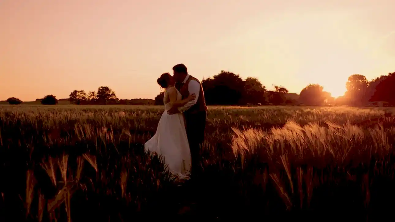 a man and woman kissing in a field of wheat at sunset as they pose for their wedding videographer on their wedding day