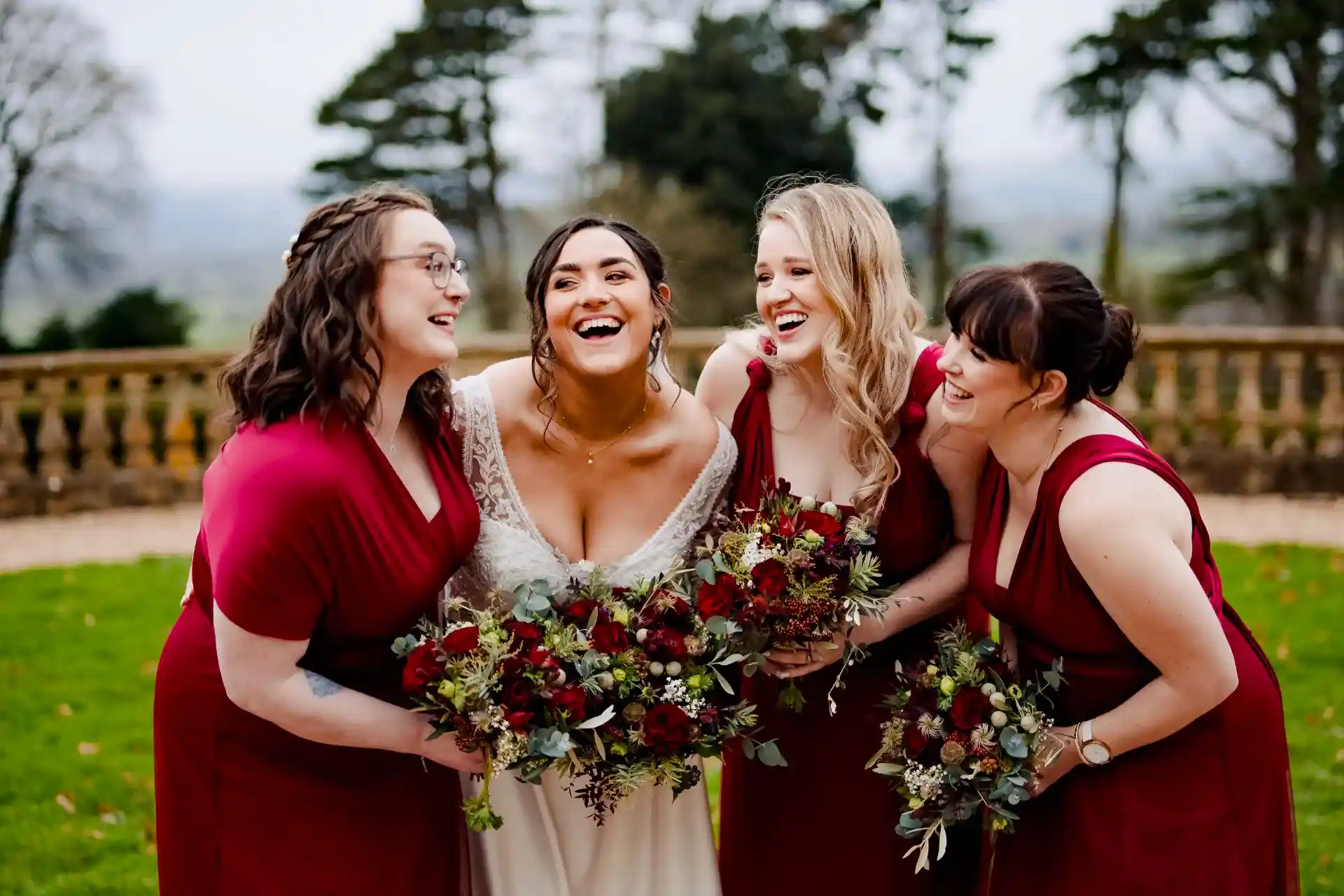 bride and her three bridesmaids who are dressed in red and holding winter boquets laugh at one another