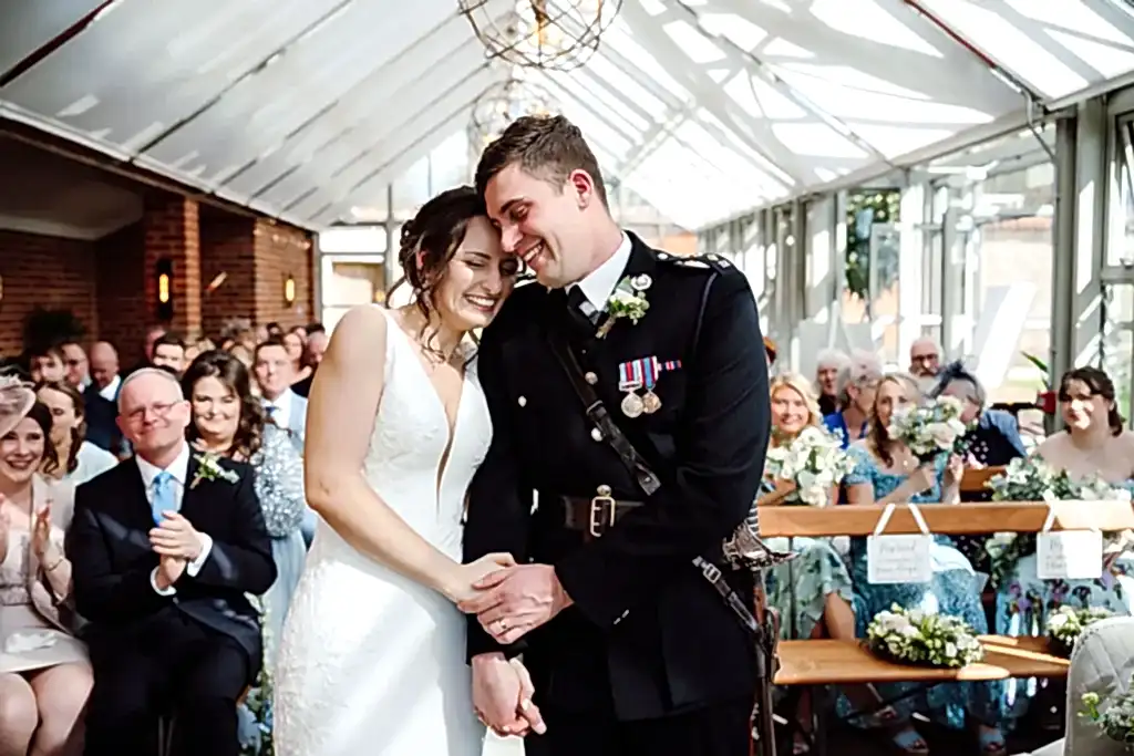 SHA0324-LOWRES-0445 bride and her husband dressed in ceremonial military uniform huddle in their wedding ceremony inside the glasshouse at Syrencot wedding venue during their wedding live stream