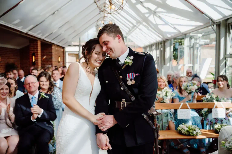 bride and her husband dressed in ceremonial military uniform huddle in their wedding ceremony inside the glasshouse at Syrencot wedding venue during their wedding live stream