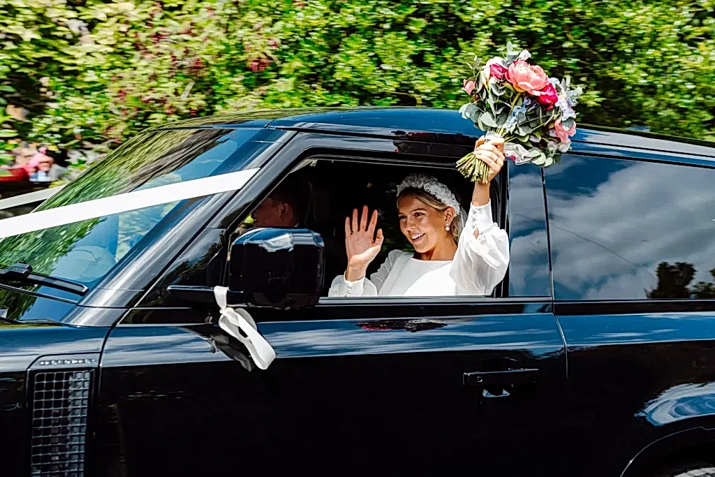 SNO0723-LOWRES-0471 bride waves out of window of black range rover holding her bouquet up in the air with her other hand