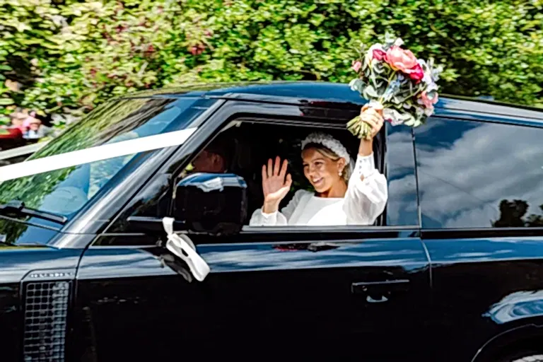 bride waves out of window of black range rover holding her bouquet up in the air with her other hand