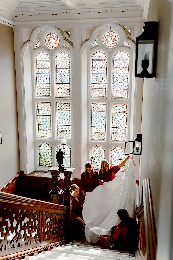 four bridesmaids in red dresses surround the bride's dress as they hang and arrange it on a light fitting in the grand staircase of Clevedon Hall wedding venue